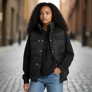 Woman wearing a black puffer vest over a black shirt with blue jeans on a blurred street background